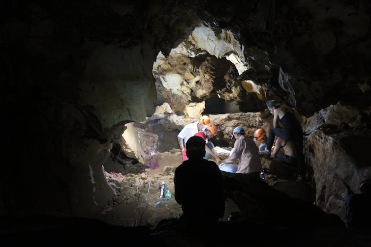 A dark cave with light walls, with several people in hard hats looking through the sediment