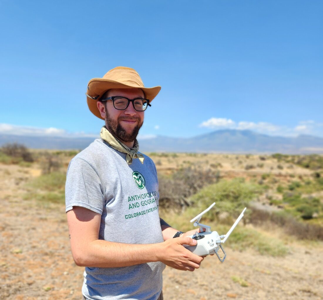 A man wearing a hat is standing in a dry landscape holding a drone controller and smiling.