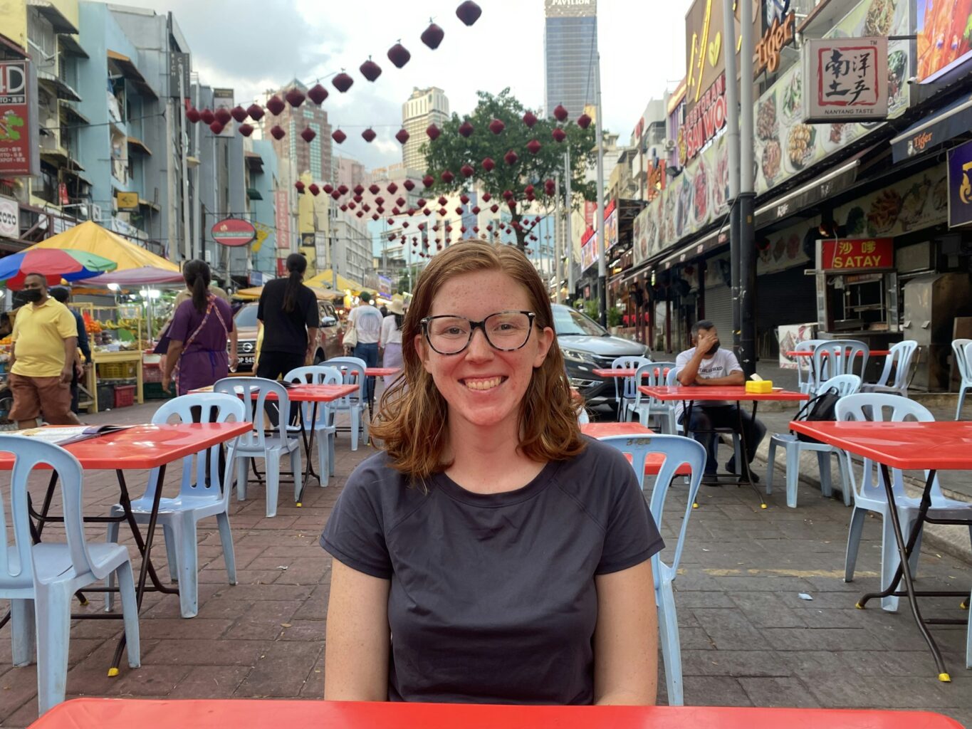 Audrey Arner, who is studying evolutionary mismatch, is sitting at a red table in an outdoor eating area on tan urban street. She has shoulder-length hair and glasses and is smiling at the camera.