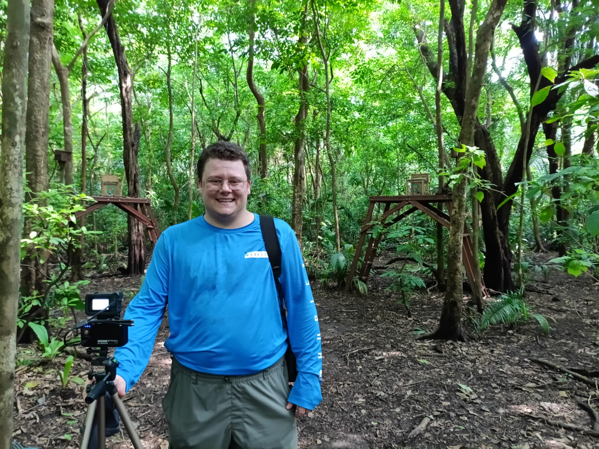 Evan Cunningham in Costa Rica. He's wearing a blue shirt and green trees are in the background. He's setting up experiment platforms for studying capuchin behavior.