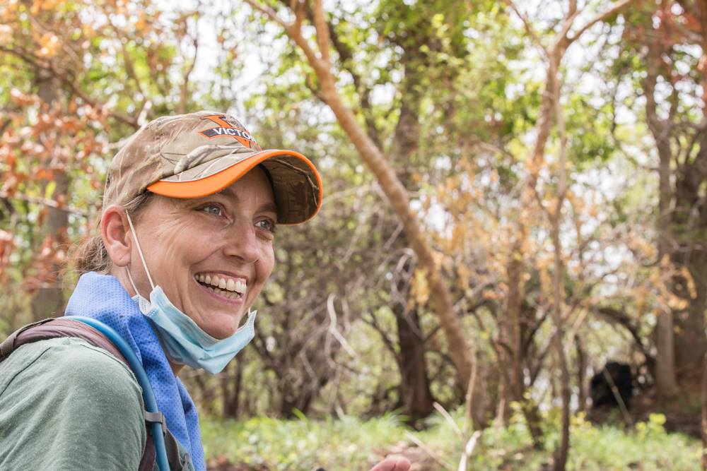 A woman in a baseball cap standing in the forest smiling. A chimpanzee is in the distance, sitting at the base of a tree.